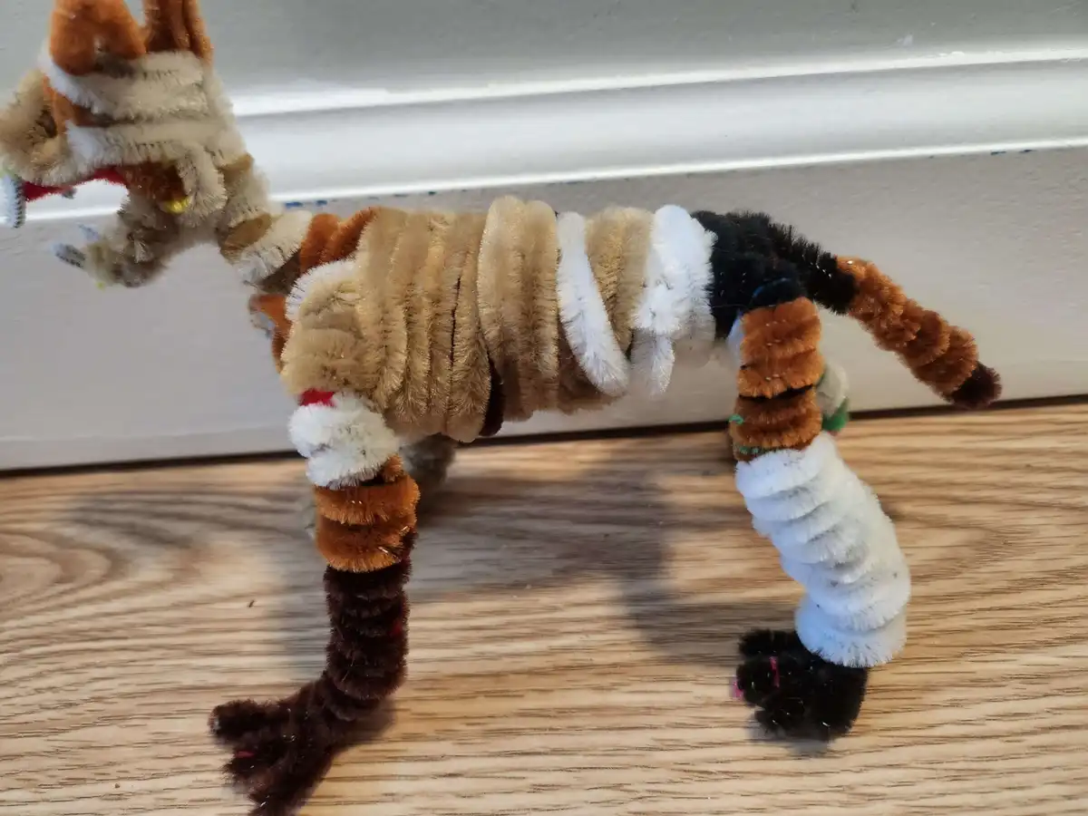 A handmade tiger figurine crafted from fuzzy pipe cleaners stands on a wooden floor, its body twisted unnaturally at the…