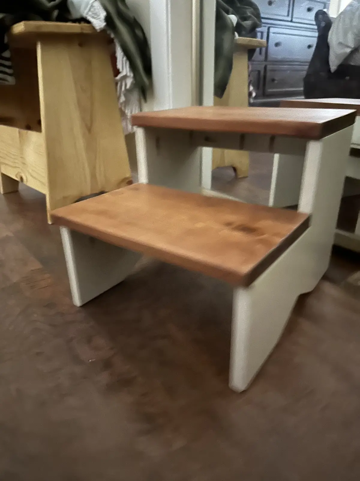 A two-step wooden stool with light-colored painted legs and natural wood tops sits on a hardwood floor, with a mirror…