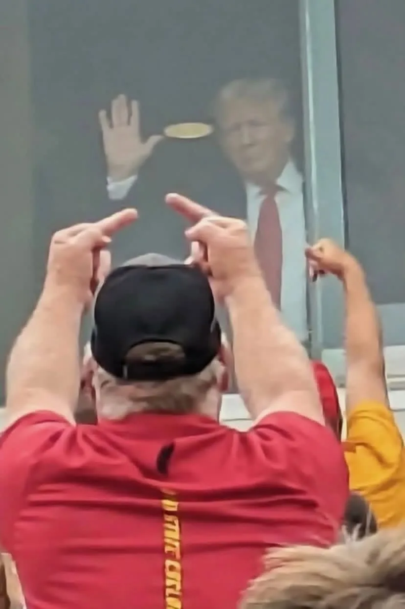 A man in a red shirt and black cap is seen from behind, pointing at a glass window where a blurry image of Donald Trump is…