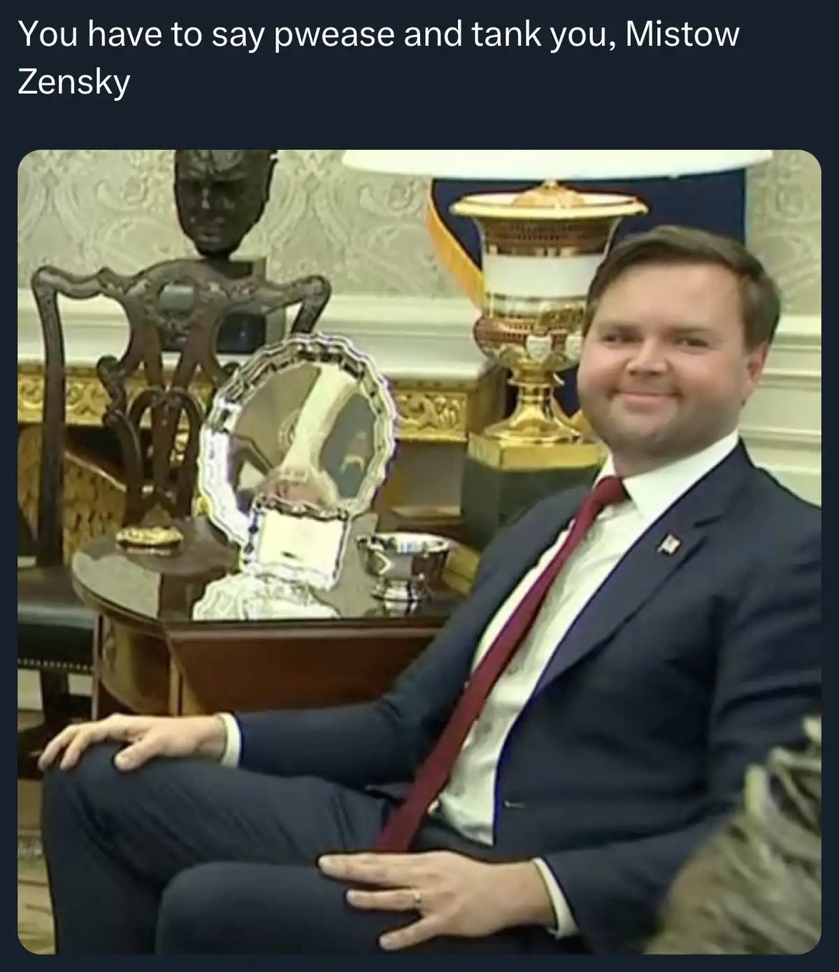 A man in a dark suit and red tie sits confidently in an ornate room with a mirror, trophy, and decorative lamp behind him…