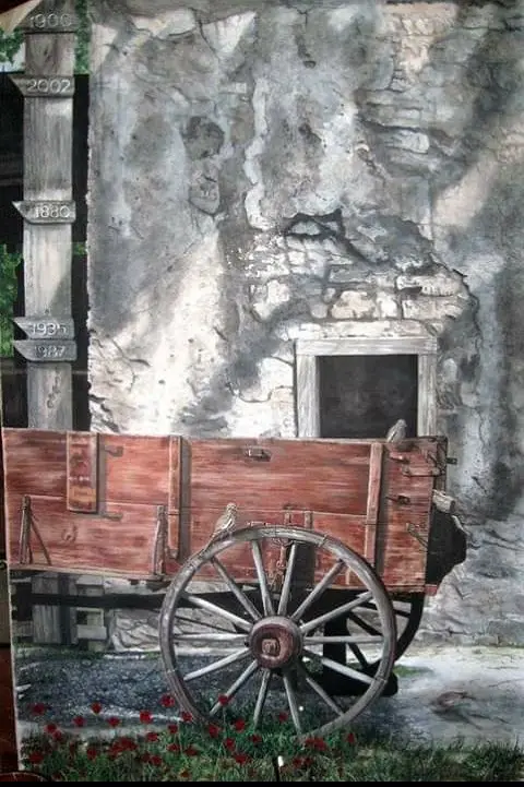 A weathered wooden cart with a large spoked wheel sits in front of a crumbling stone wall. A vertical wooden post to the…