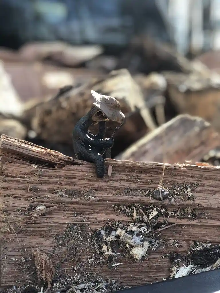 A small, dark figurine of a person wearing a conical hat sits on a rough, splintered piece of wood, holding a stick. A…