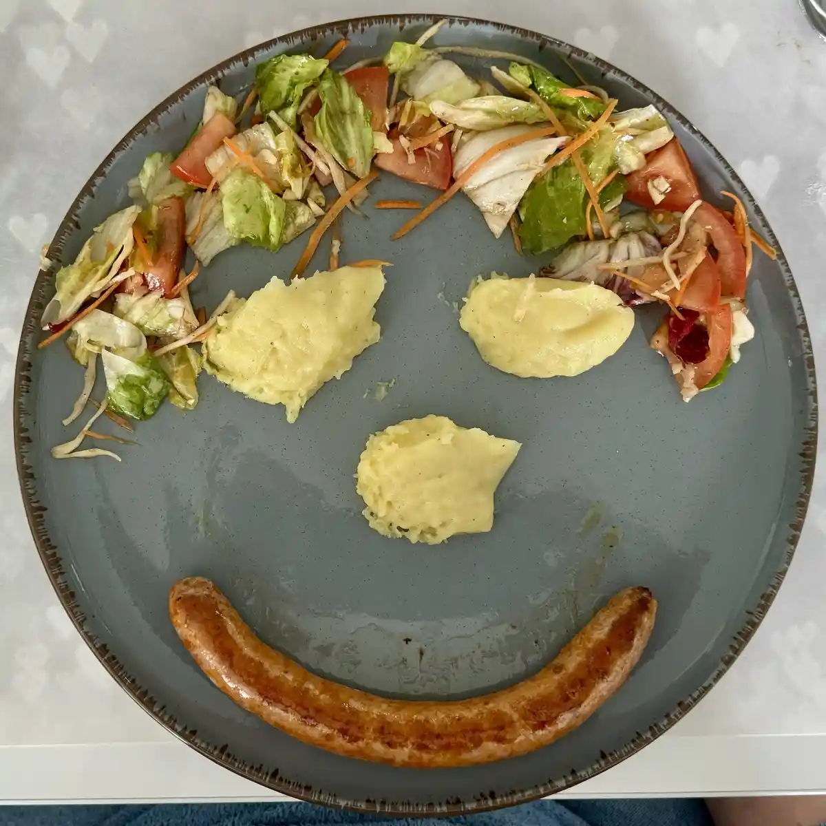 A plate of food arranged to resemble a smiley face, with salad forming the eyes and eyebrows, three lumps of mashed potatoes…