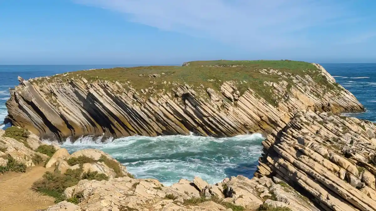 A scenic photo of layered, tilted rock formations along a coastline with turquoise ocean waves crashing against them under a…