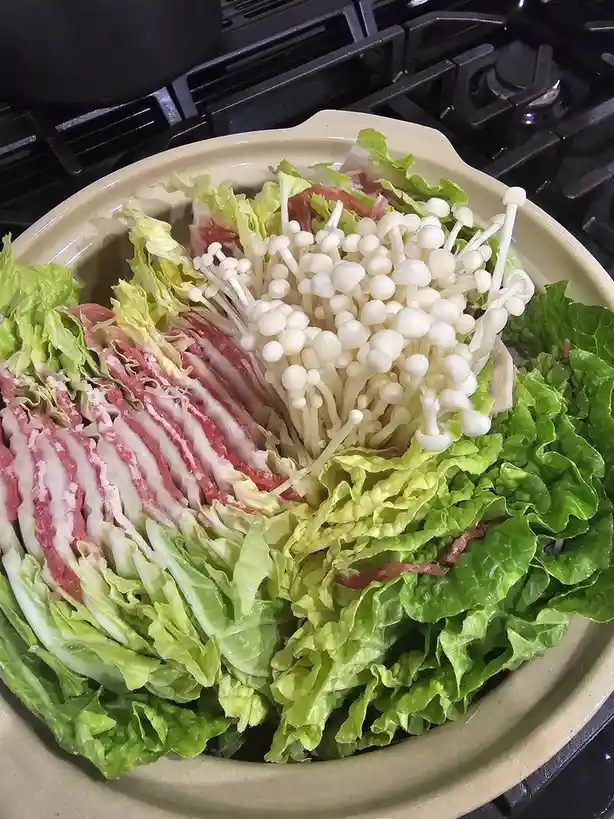 A beige ceramic hot pot sits on a black stovetop, filled with neatly arranged ingredients including green lettuce leaves…