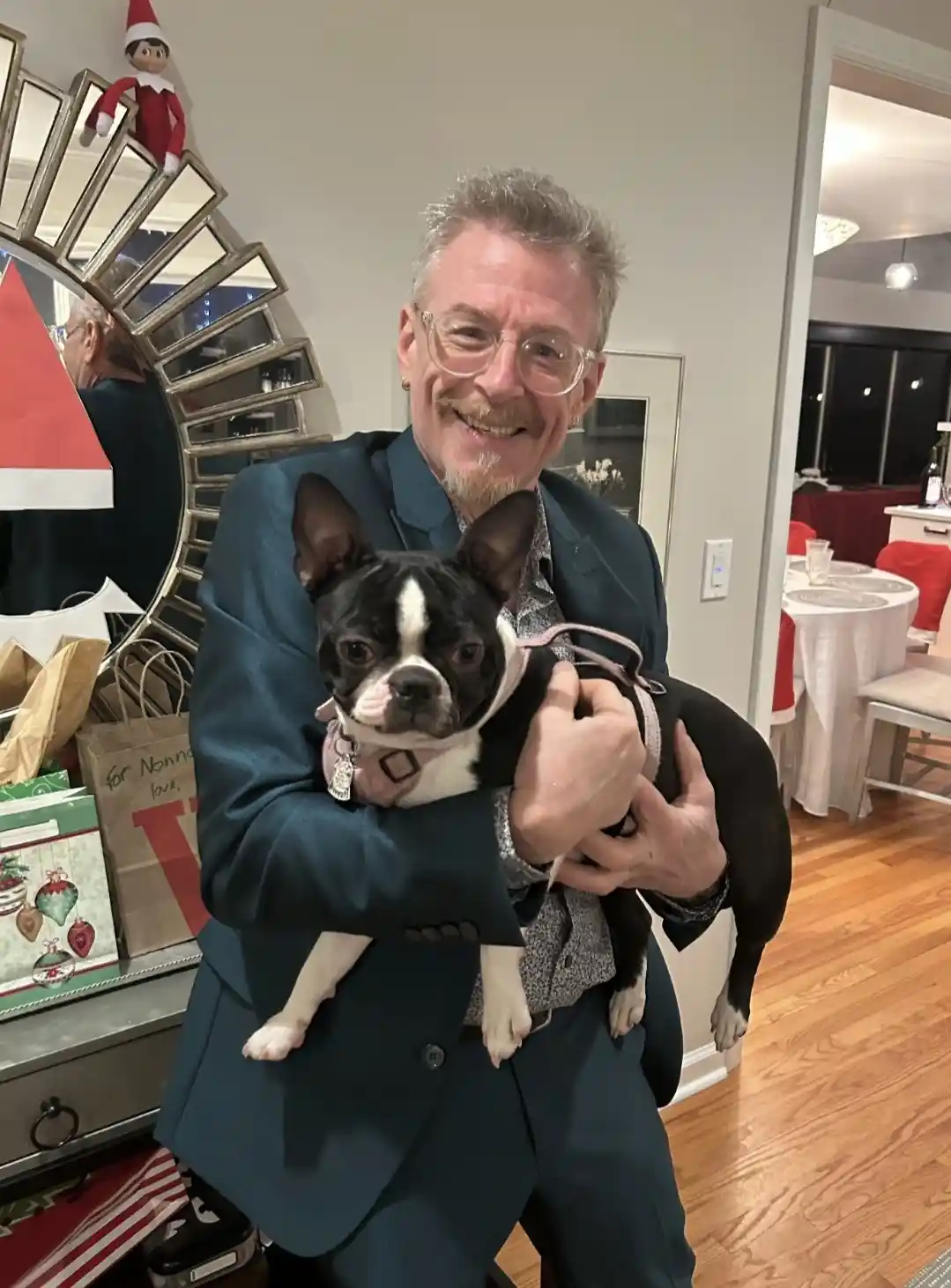 A smiling man in a dark blue suit and glasses holds a black and white Boston Terrier dog. The dog wears a pink harness and…