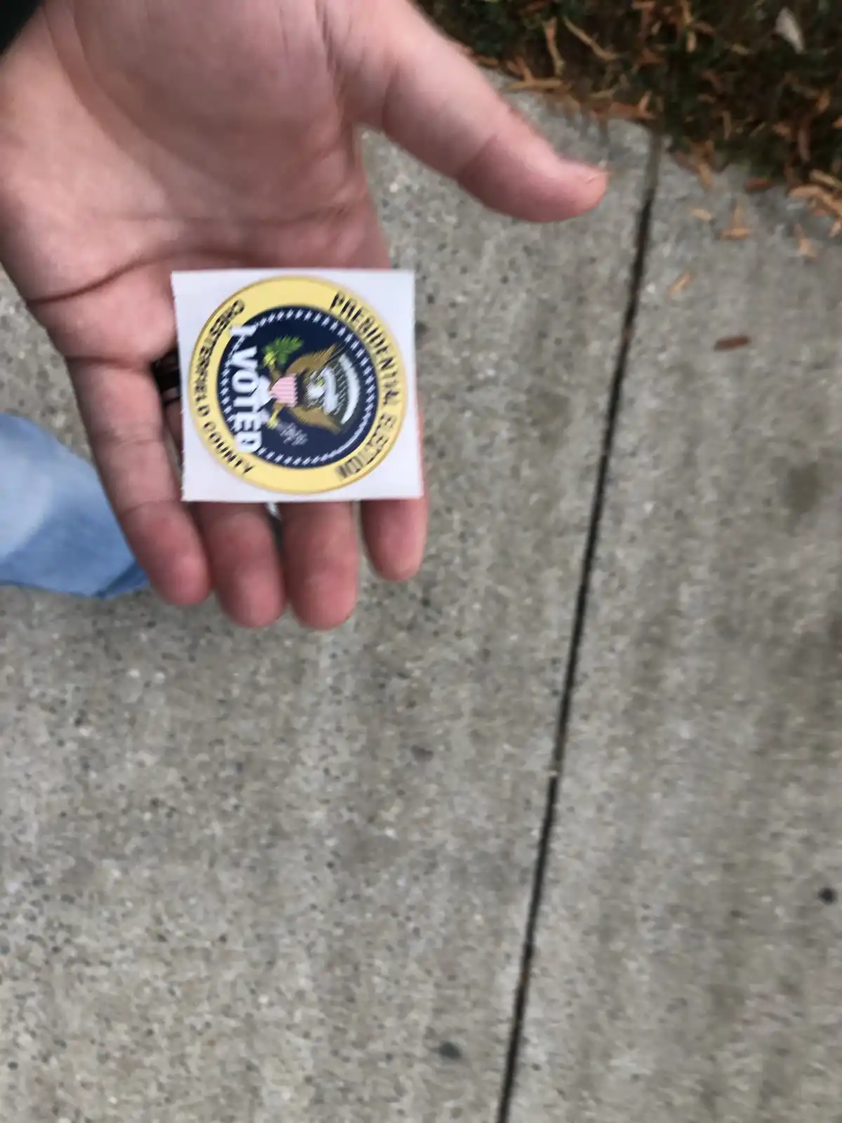 A person holds up a small, square sticker with a circular design resembling the Presidential Seal, featuring the text 'I…