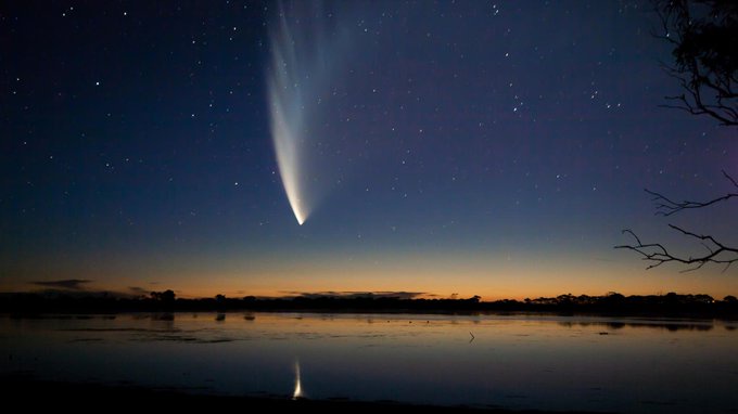 A night sky photograph shows a bright comet with a long, glowing tail arcing over a calm body of water reflecting the stars…