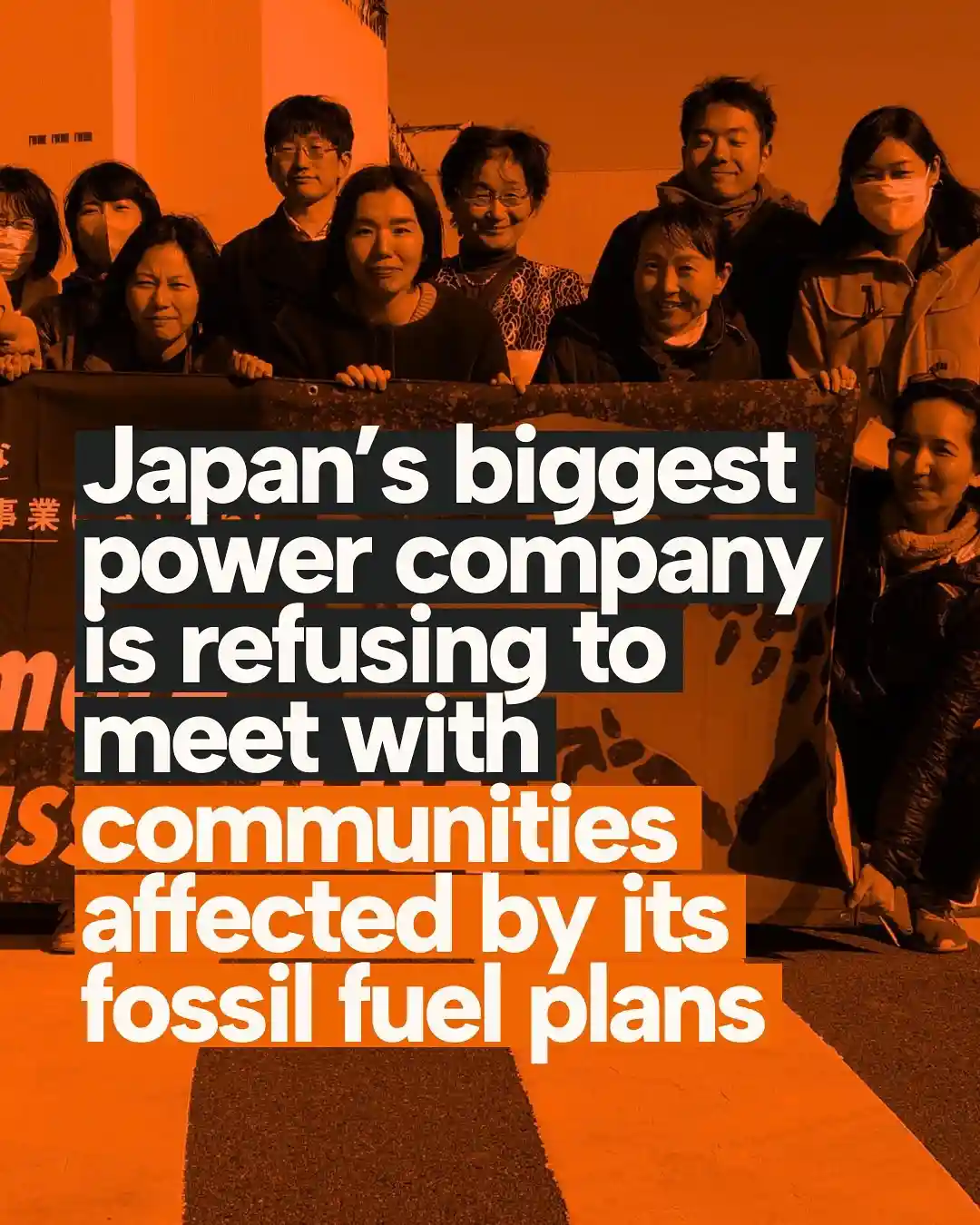 A group of Japanese people, some wearing masks, stand together holding a banner in an orange-tinted photo. Overlaid white…