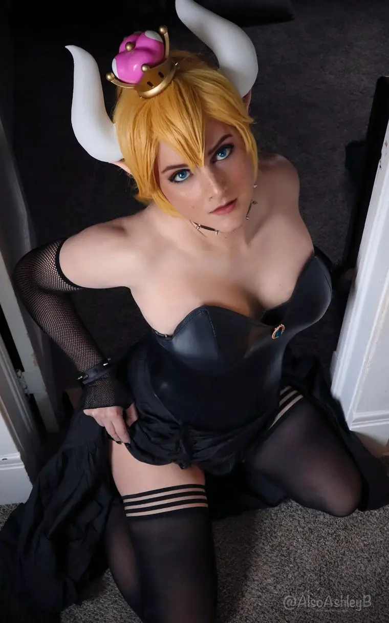 A person in cosplay as a video game character with blonde hair, white horns, and a pink crown sits on the floor looking up…