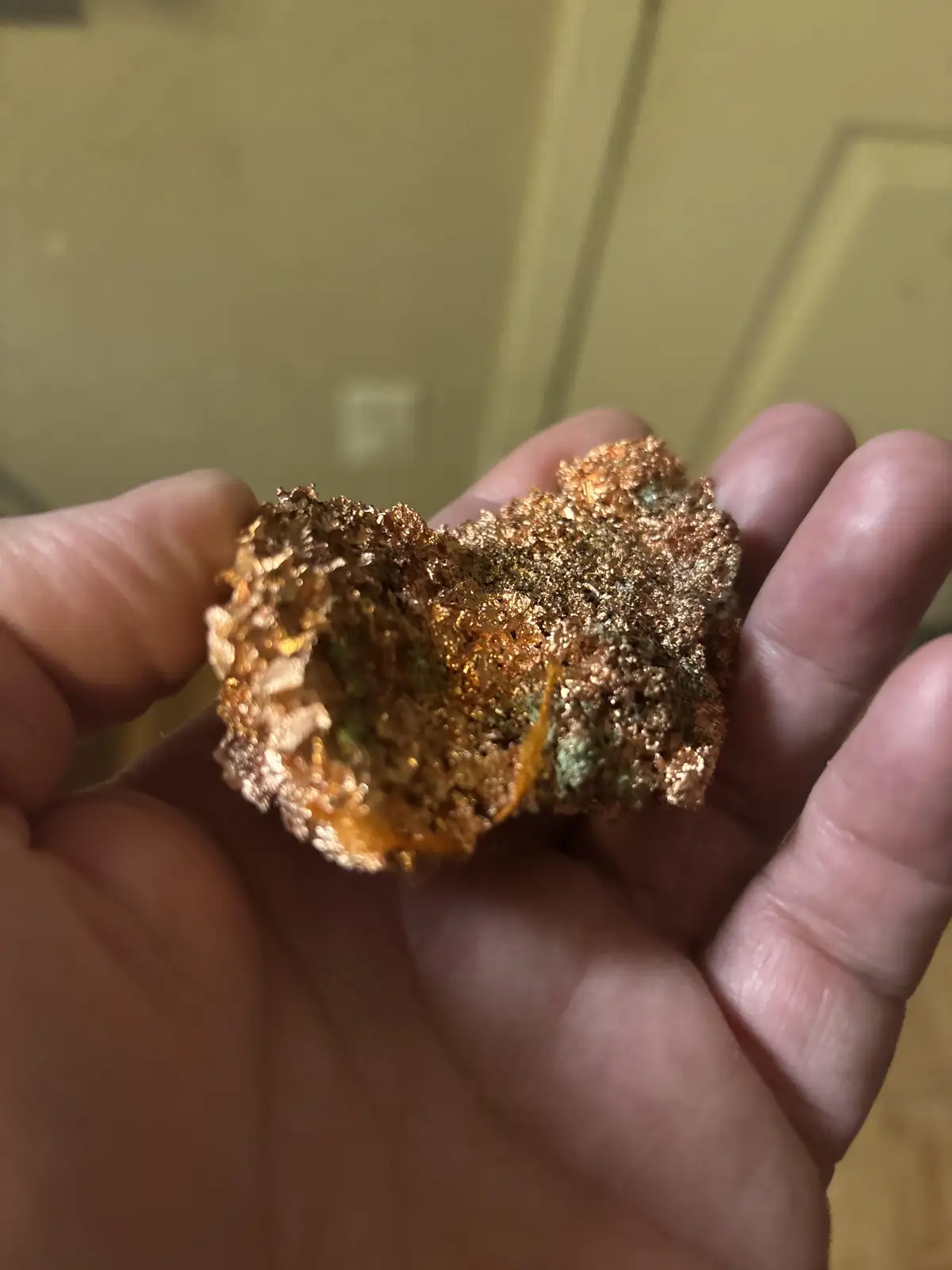 A hand holds a rough, irregularly shaped mineral specimen with a shiny, metallic copper-colored surface and some greenish…