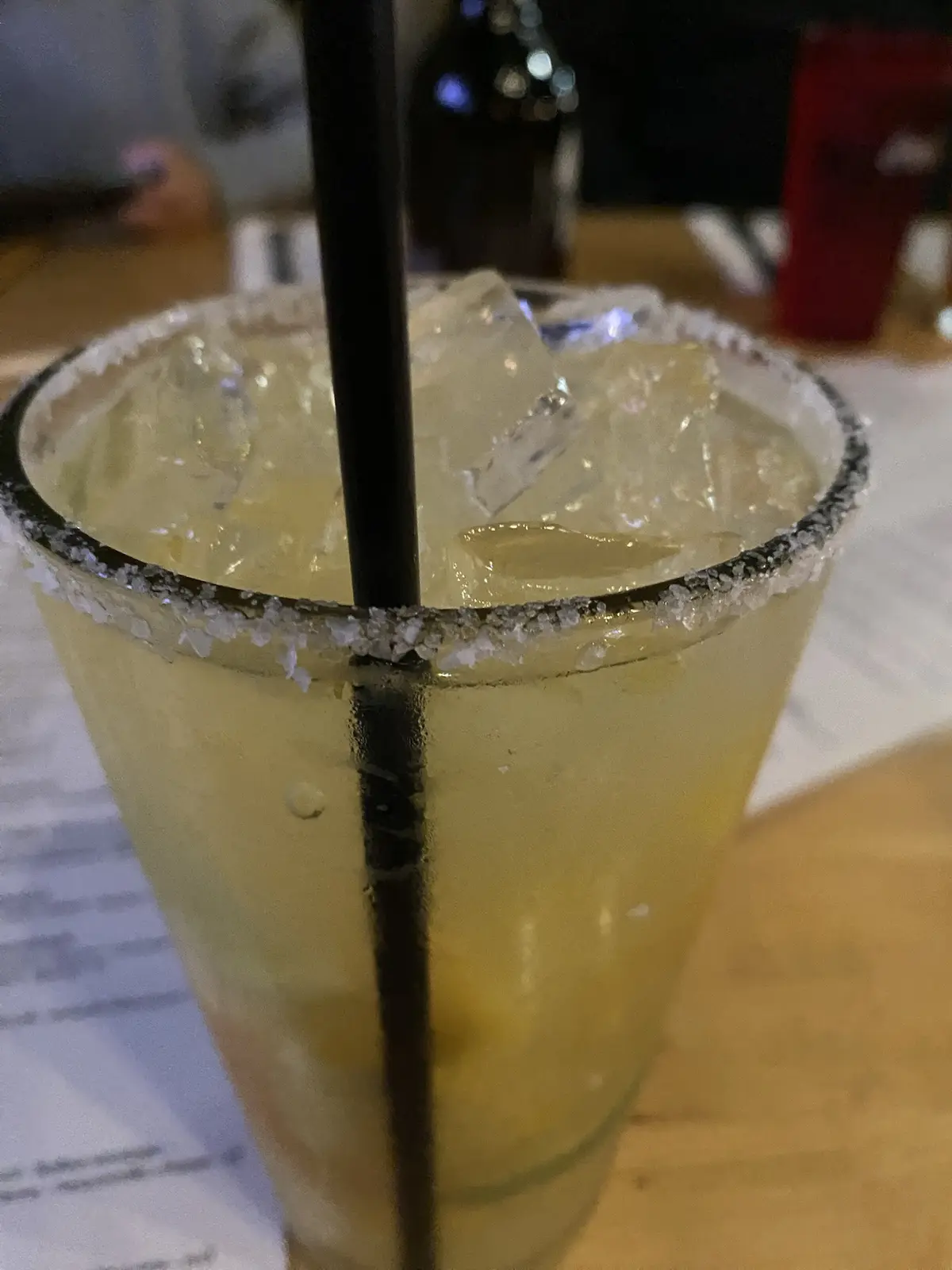 A close-up photo of a margarita in a glass with a salted rim, filled with ice and a black straw, sitting on a wooden table…