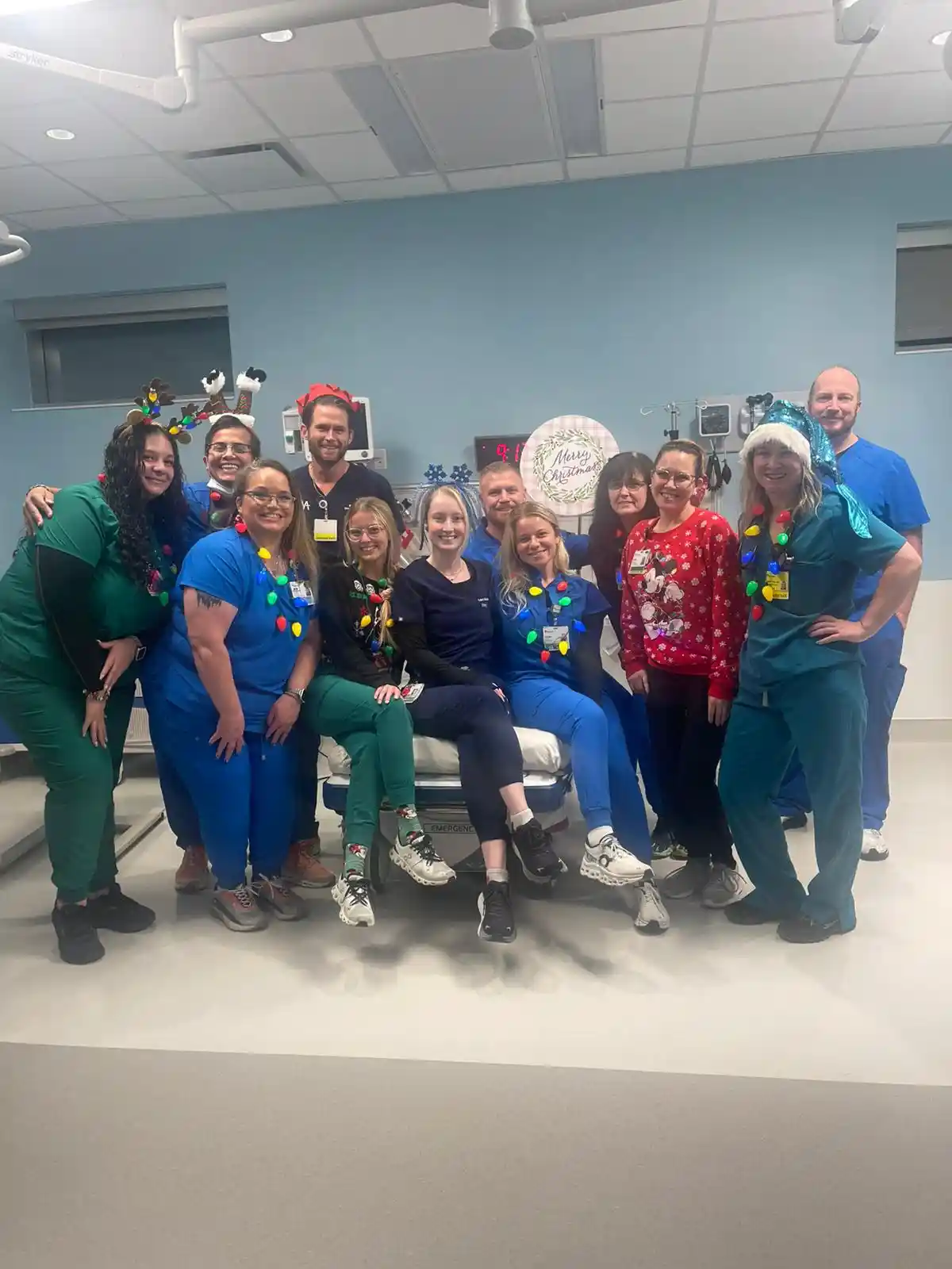 A group of medical staff in scrubs pose for a photo in a hospital room decorated for Christmas, wearing festive accessories…