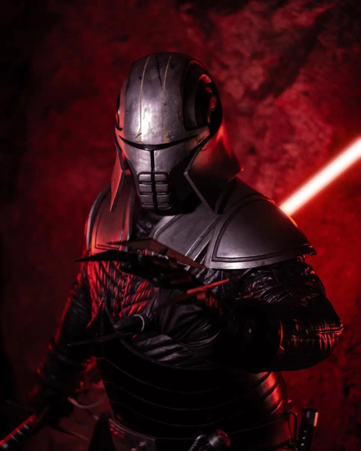 A person in a detailed, dark metallic helmet and armor stands in a red-lit, smoky environment, holding multiple blades. A…