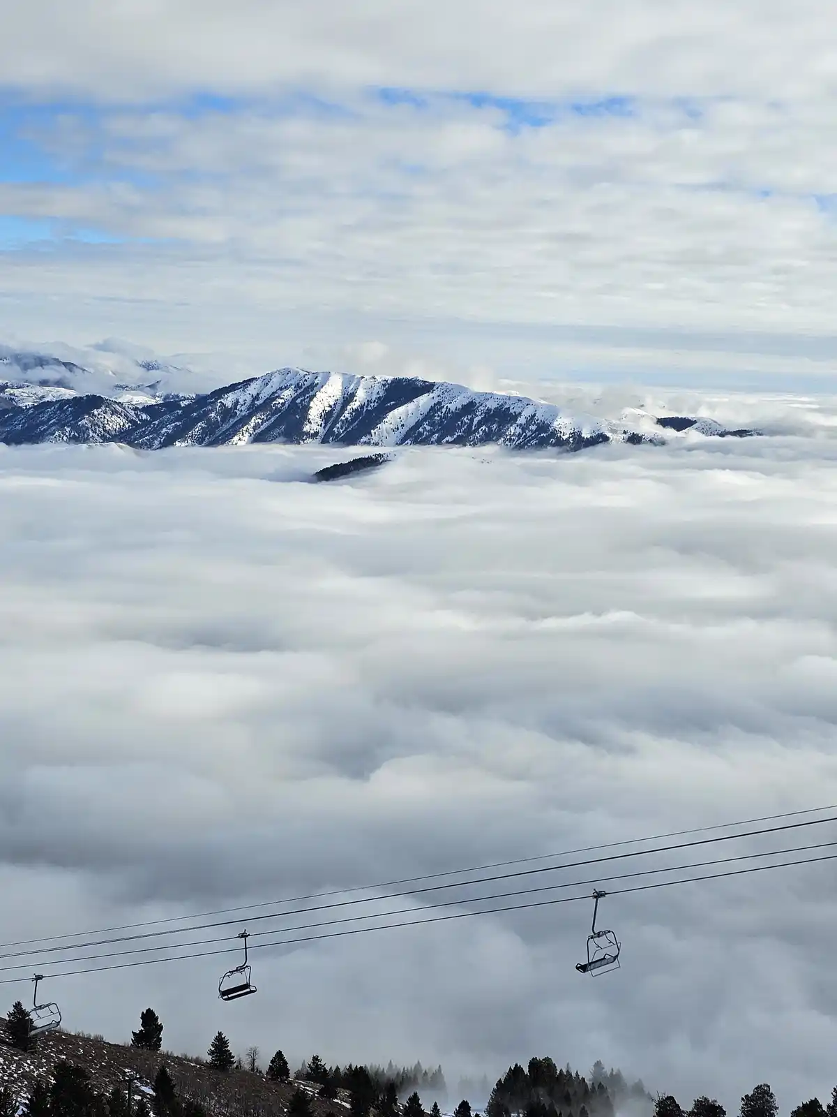 A scenic photo shows snow-capped mountains rising above a thick blanket of clouds, with empty ski lift chairs suspended on…