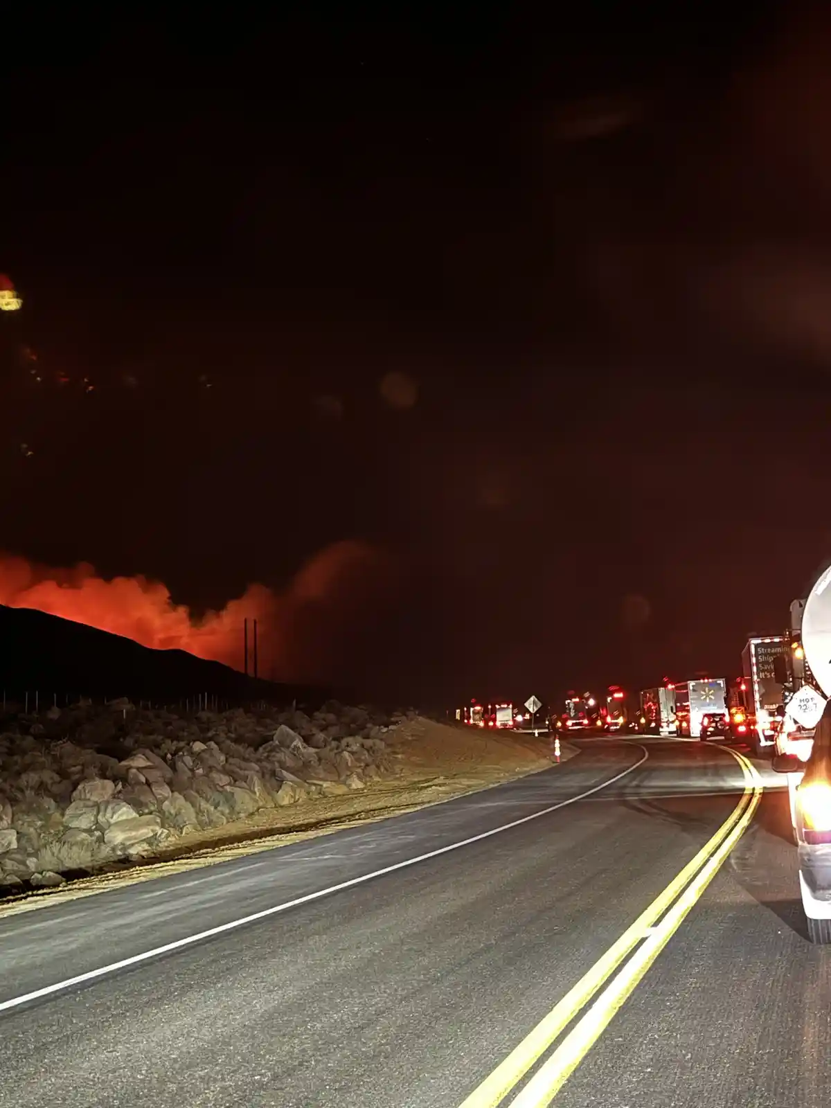 A nighttime highway scene shows a large wildfire burning on a hillside to the left, casting an orange glow across the dark…