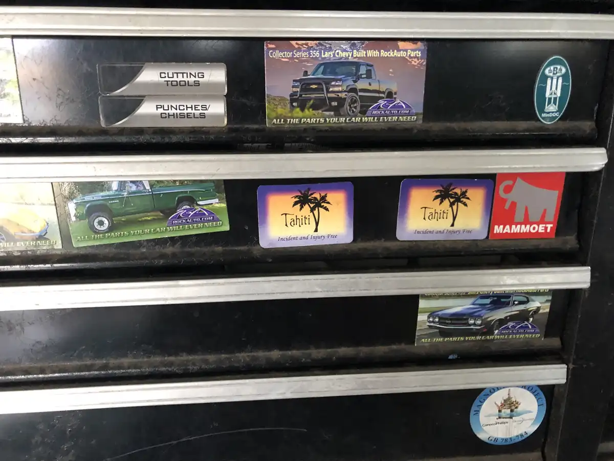 A close-up of a black tool chest with silver handles, covered in various stickers. Several stickers advertise 'RockAuto.com'…