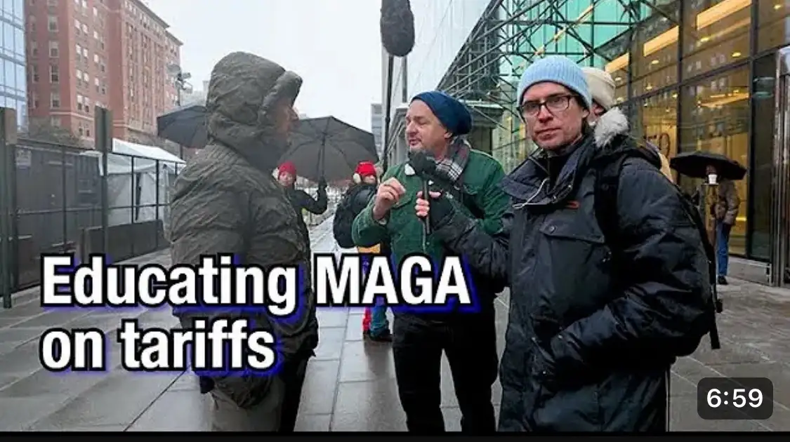 A man in a green jacket holds a microphone while speaking to another person in a hooded jacket on a rainy city sidewalk. A…