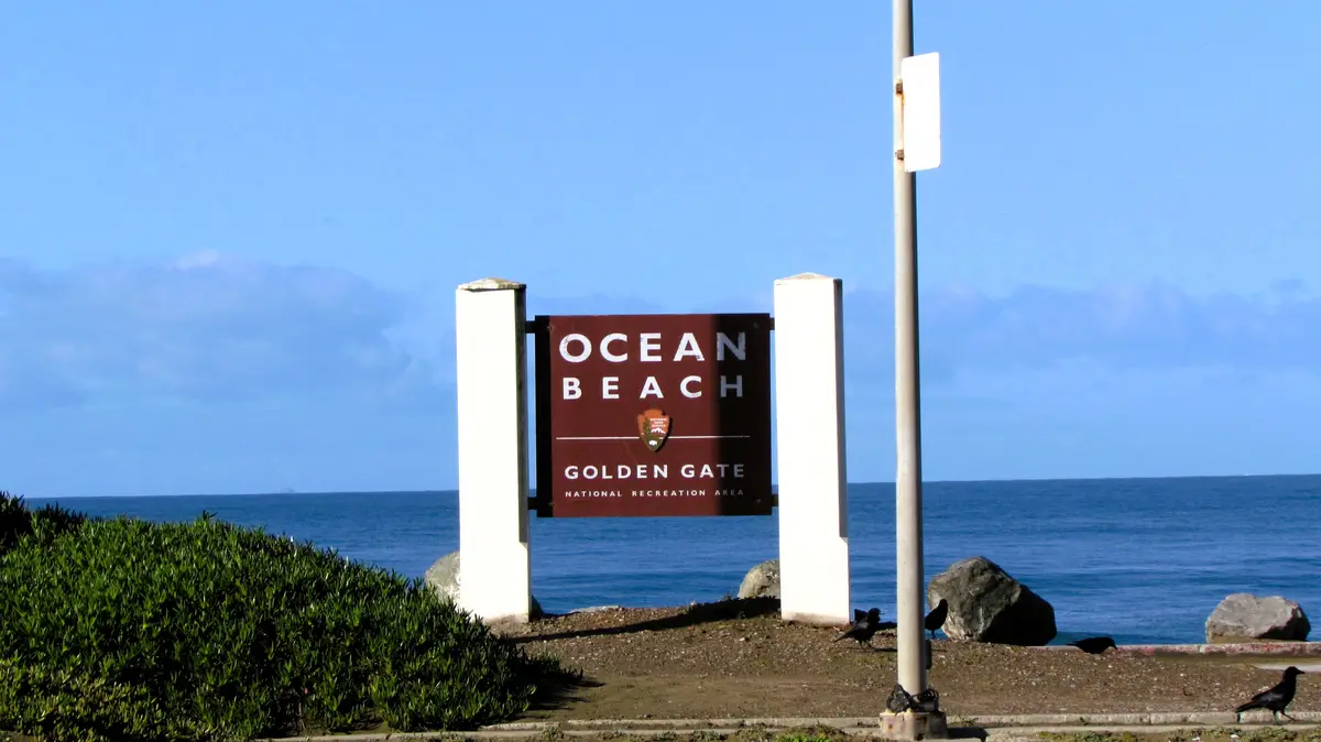 A brown sign reading 'OCEAN BEACH GOLDEN GATE NATIONAL RECREATION AREA' is mounted between two white pillars near a beach…