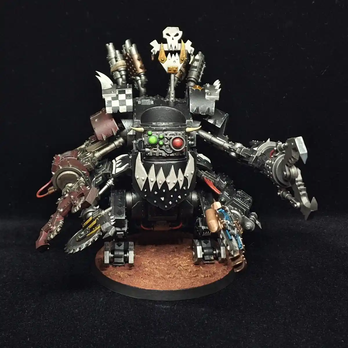 A heavily armored, multi-limbed, menacing robotic figure with glowing red and green eyes, sharp teeth, and various weapons…