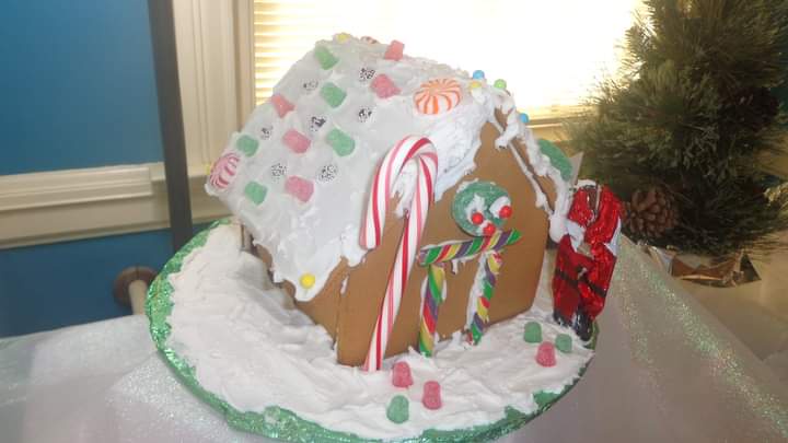 A decorated gingerbread house with candy canes, gumdrops, and icing sits on a snowy base, with a small Santa Claus figurine…