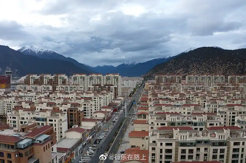一张从高处俯瞰的城市全景照片，画面中密集排列着大量米白色外墙、红棕色屋顶的现代住宅楼，一条宽阔的道路贯穿其中，远处是连绵起伏、部分山顶覆盖着积雪的山脉，天空布满厚重的云层。图片右下角有微博水印“@衍乐在路上”。