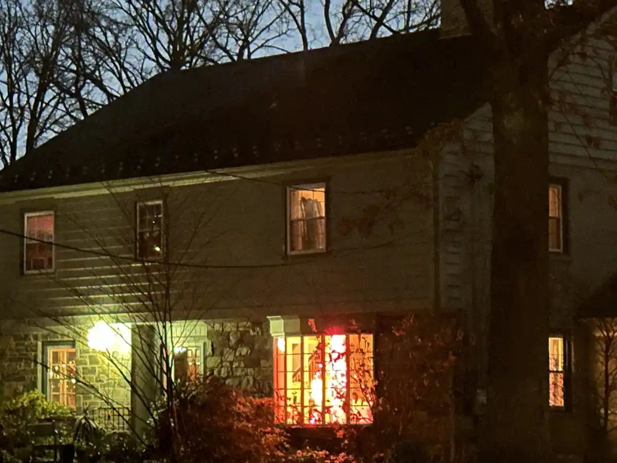 A nighttime photo of a two-story house with lit windows, one of which glows intensely red, contrasting with the dimmer…